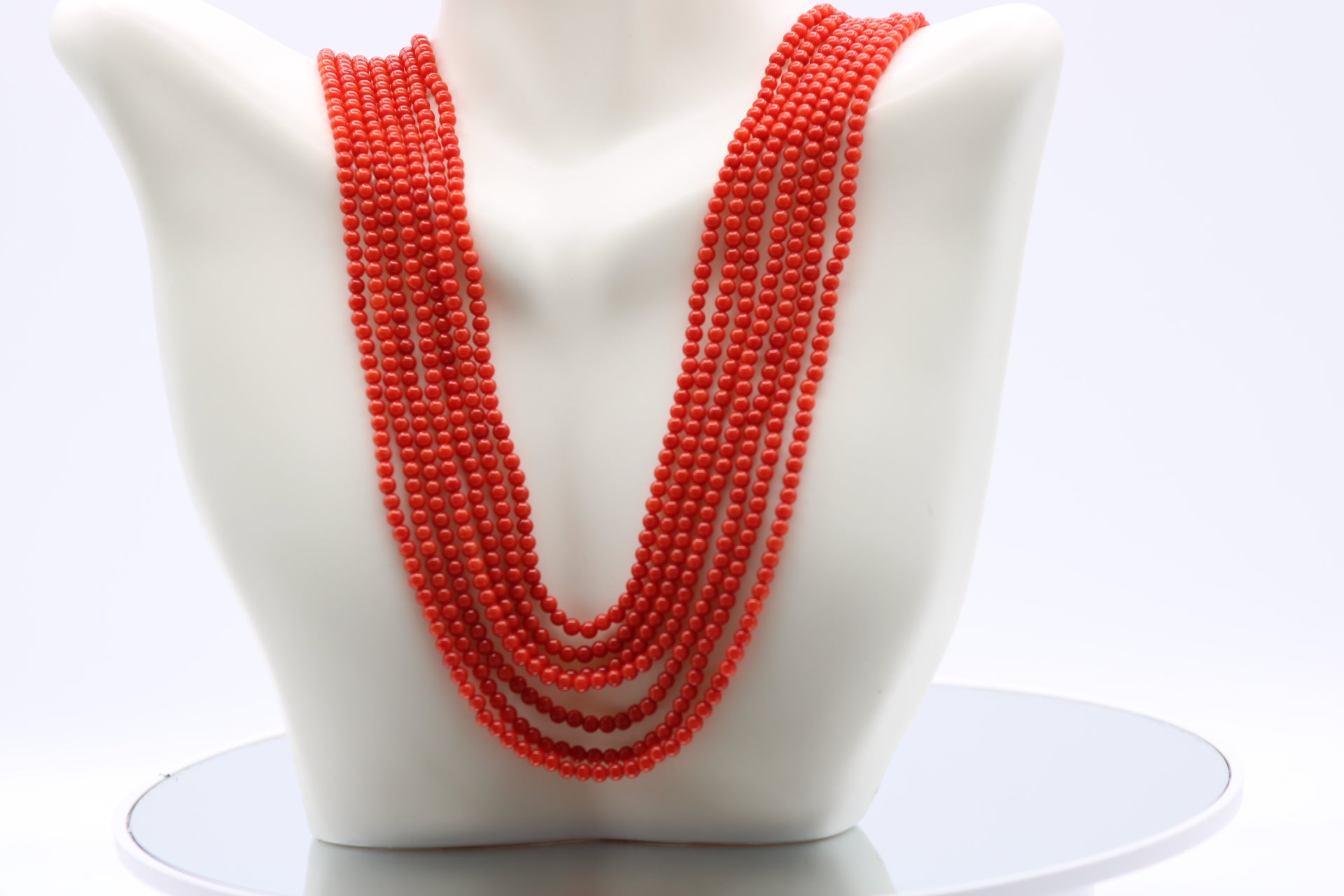 Coral necklace 3mm with 7 strands in reddish orange color displayed on a bust form.